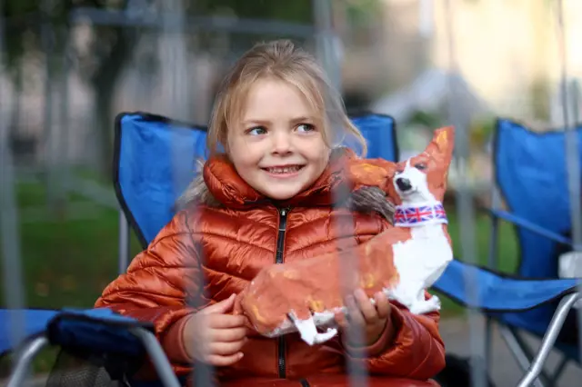 A girl holds a corgi as people wait for the coffin