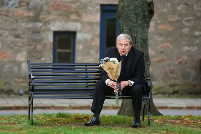 A man sits on a bench while waiting for the procession near Balmoral