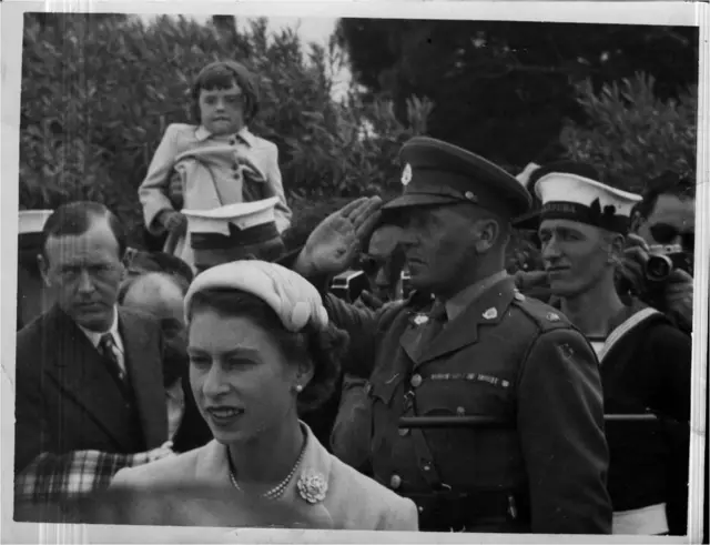 A young Janine is hoisted up above over her father's head to see the Queen in Malta over the heads of a crowd