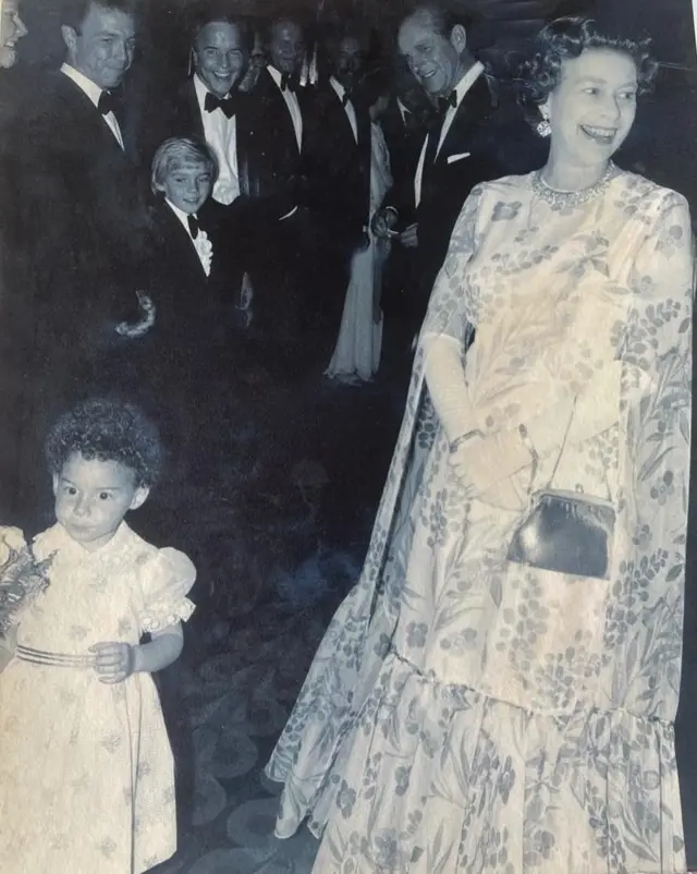 The Queen laughing as  toddler stands holding a bunch of flowers