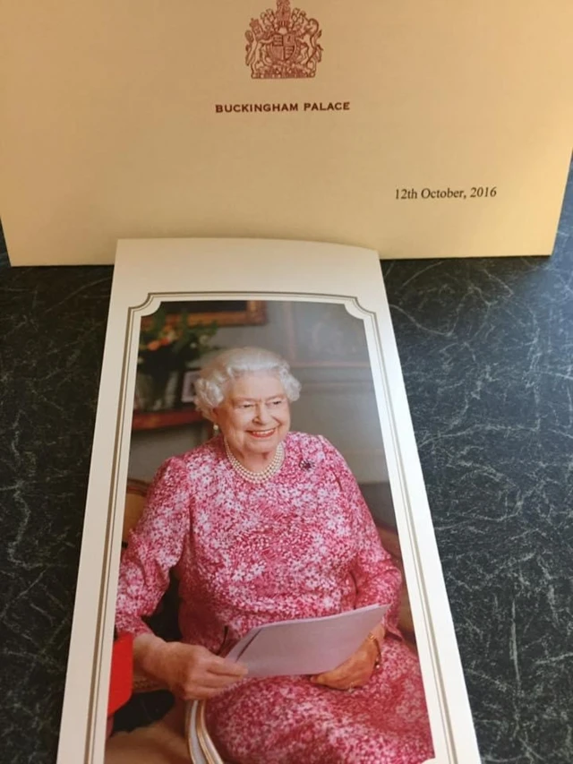 A card with a photograph of the Queen in a red patterned dress holding documents