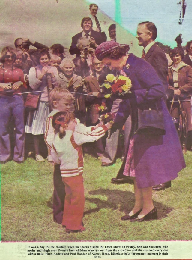 The Queen accepts flowers from two small children, she is wearing a purple tea dress and a long purple coat with a matching purple hat