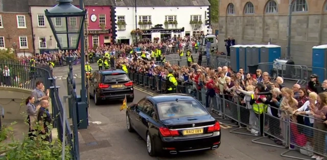 Crowds of people behind barriers wave at two cars driving away