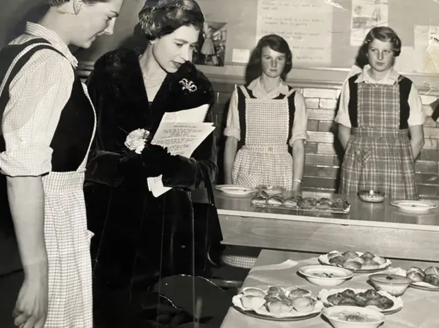 The Queen looks at cakes and pastries made by the girls of Barnet school, behind her stands two girls in aprons, one is Lynda Carter