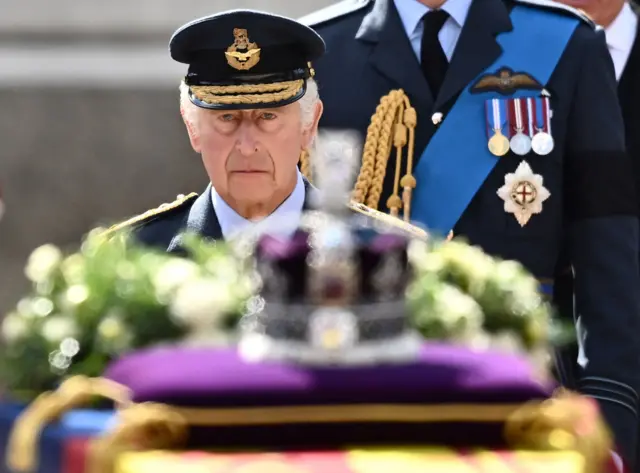 King Charles III walks behind the coffin of Queen Elizabeth II, which is adorned with a Royal Standard and the Imperial State Crown