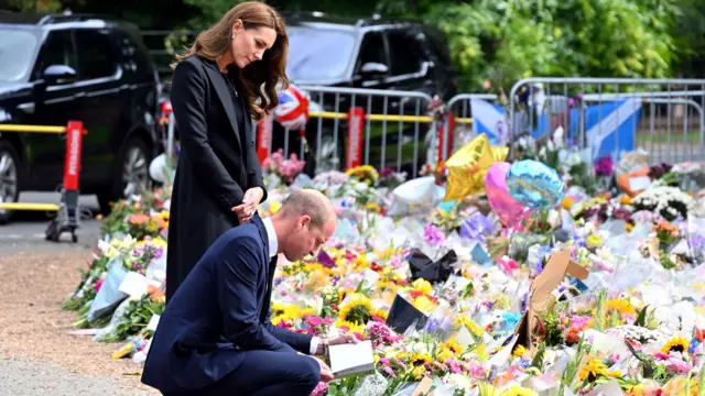 William, the Prince of Wales, bends down to read tributes left outside Sandringham, while Kate, the Princess of Wales, stands above him