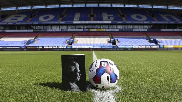 Queen on cover of football programme next to ball in stadium