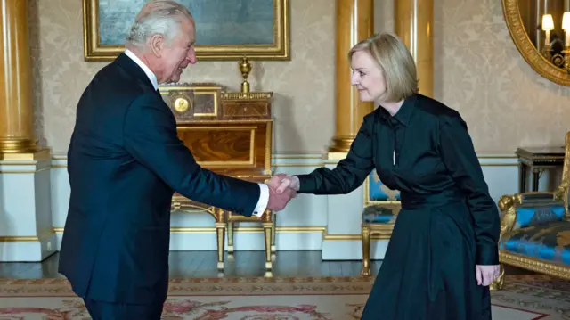 King Charles III, wearing a black suit, shakes hands with UK PM Liz Truss, wearing a long black dress, during their meeting at Buckingham Palace