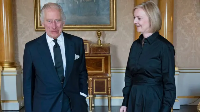 King Charles III, wearing a black suit, hosts UK PM Liz Truss, wearing a long black dress, at Buckingham Palace