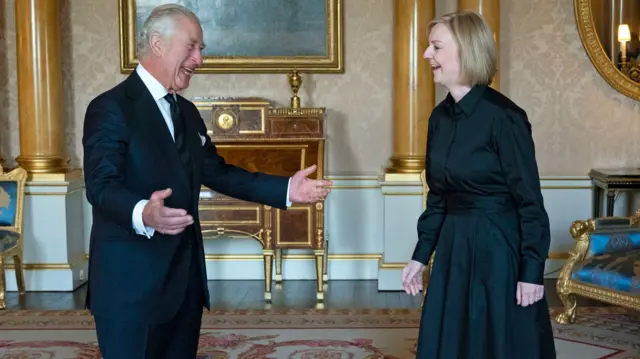 King Charles III, wearing a black suit, gestures to UK PM Liz Truss, wearing a long black dress, during their meeting at Buckingham Palace