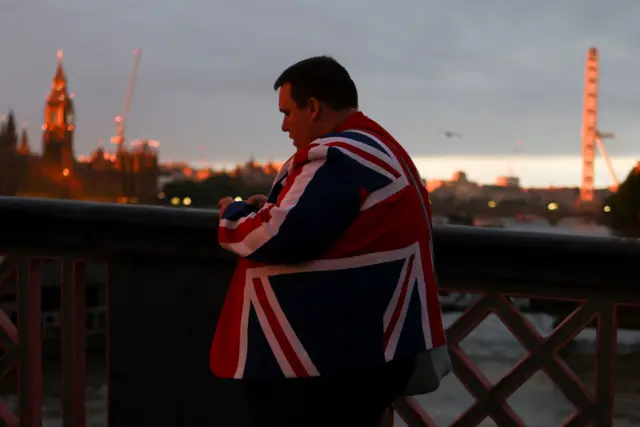 A person wearing a suit with a UK flag pattern queues in the dusk light