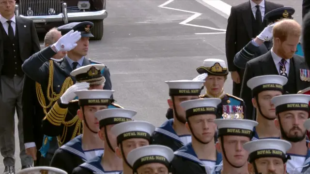 Image shows Prince William giving a salute, with King Charles doing the same in front of him and Princess Anne to his left also doing so