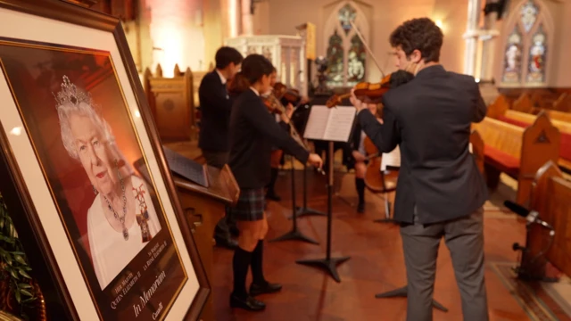 A college orchestra rehearses next to a mounted picture of the Queen