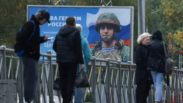 People gather at a tram stop in front of a board displaying a portrait of Russian service member Sergei Tserkovniy in Saint Petersburg,