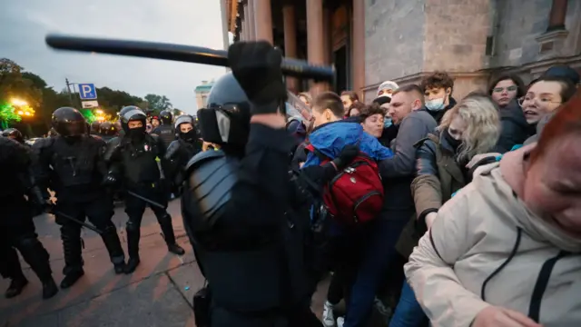 A Russian police officer waves his baton at a group of people involved in the protest, with one woman (front right) closing her eyes and appearing to cry