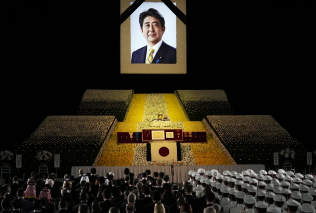 Packed rows of guests stand for a minute silence in front of a portrait of Mr Abe