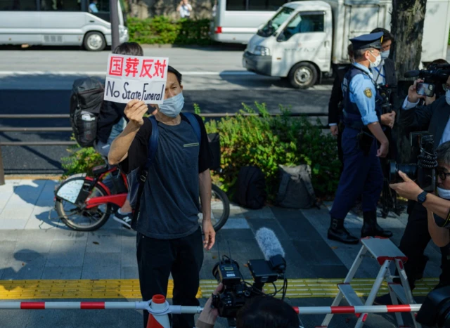 A protester holds a sign reading 'No State Funeral' outside the funeral venue this morning