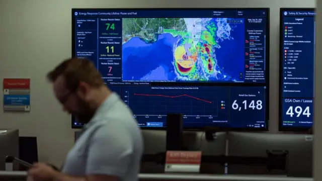 A FEMA agent glances at his phone in front of a board mapping the devastation caused by Hurricane Ian