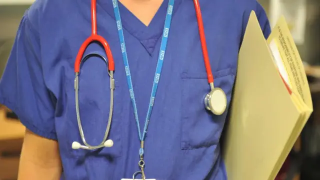 Generic shot of the upper body of an NHS medic, who wears a stethoscope and carries a binder of information