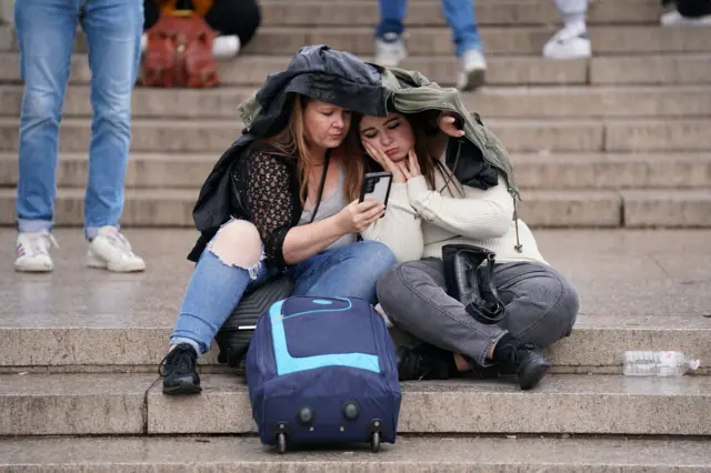 Two women sitting under a jacket on the stone steps up to The Queen Victoria Memoria, reading news updates on their mobile phone