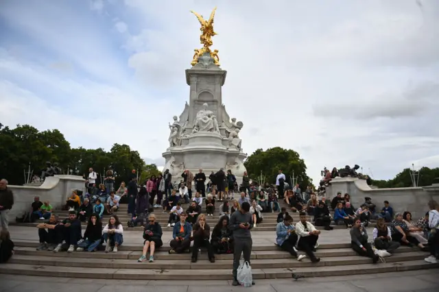 People sit on the steps of the Queen Victoria Memorial in front of Buckingham Palace