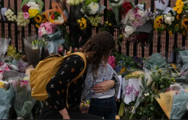A woman comforts her child as they both embrace outside the palace next to flowers