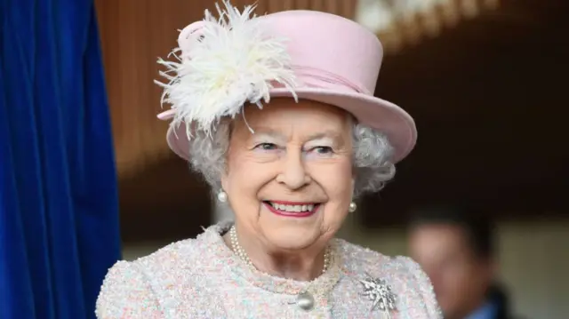 The Queen, smiling in a pink hat with white flower