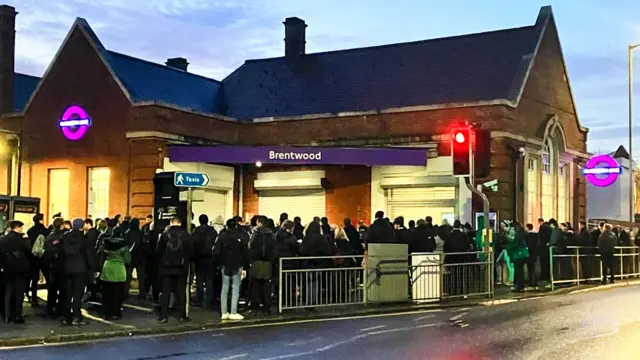 A long queue of people standing in the dark outside Brentwood station