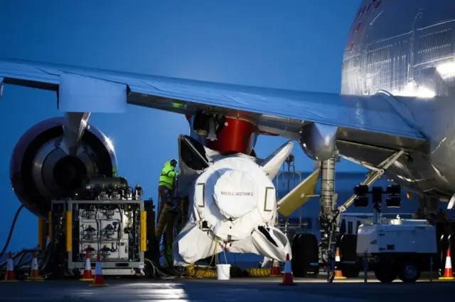 Technicians work on Virgin Orbit's LauncherOne rocket, attached to the wing of Cosmic Girl, a Boeing 747-400 aircraft.