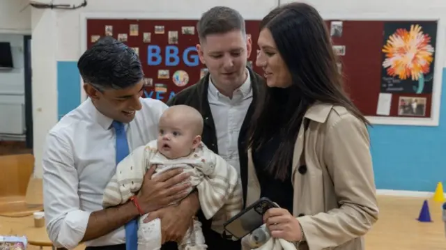 Rishi Sunak holding a baby, the child's parents are stood by him