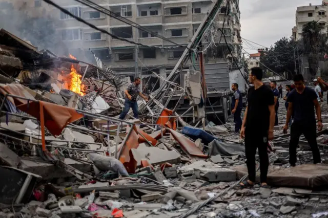 Palestinians examine the rubbles of a destroyed residential building, one man is searching it while a fire continues to burn in the background