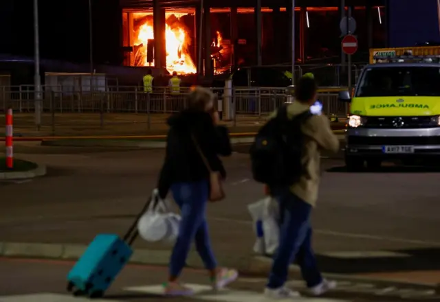 People walking with suitcases looking at a huge fire in the background