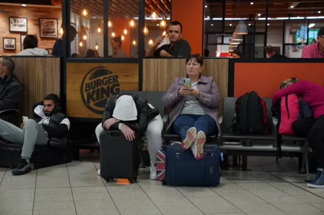 Passengers wait inside Luton Airport departures, one woman has her feet up on a suitcase while another is resting his arms and head on a suitcase.