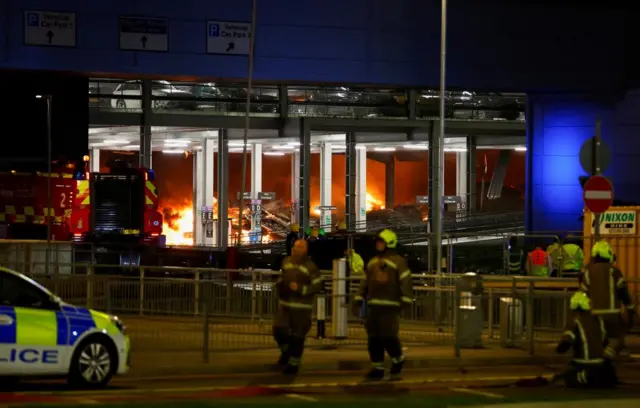 Firefighters and a police car in front of the fire in the car park