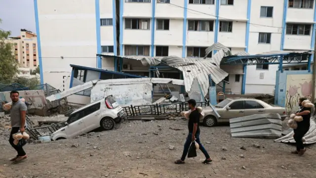 Palestinian men carrying bread walk past damaged cars and a crater in front of an Unrwa school