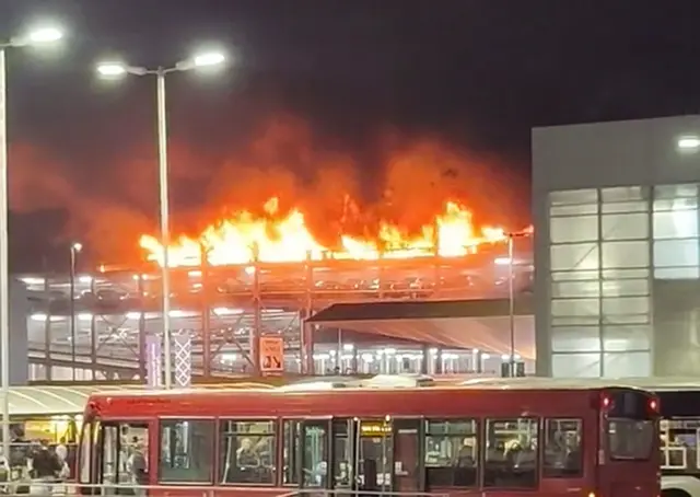 Flames and smoke billow from the car park as passengers getting off a bus look on