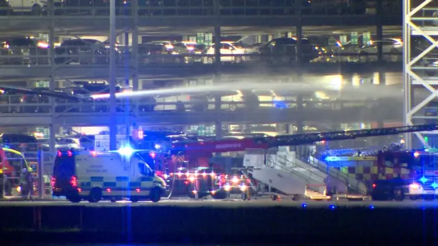 A crane, ambulance and multiple fire crews are outside a car park. Water from an appliance out of shot is spraying water.