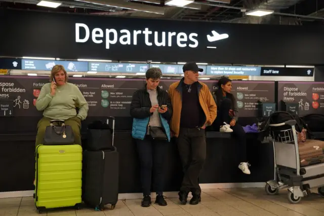 Passengers wait inside Luton Airport departures, one woman is speaking on the phone, while another is looking at her