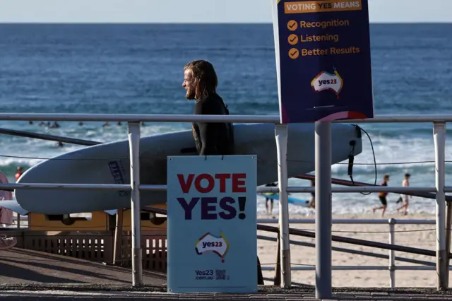 A surfer carrying his board walks past a Vote Yes sign