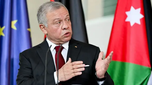 King Abdullah speaks at a podium in front of EU, German and Jordanian flags