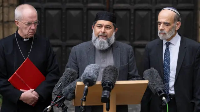 Justin Welby, the Archbishop of Canterbury, Imam Shaykh Ibrahim Mogra, and Rabi Jonathan Wittenberg give a press conference in front of microphones at a lecturn