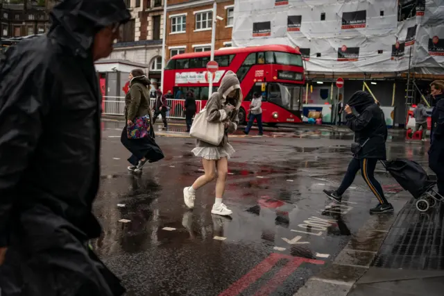 People walk through rainy streets of London with umbrellas and hoods on heads