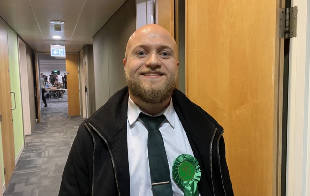 Cade Sibley, Green candidate in Mid Bedfordshire, smiling at the count. He is wearing a dark green tie, white shirt and black jacket. He also has on a green party rosette.