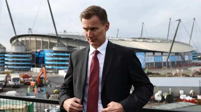 Chancellor Jeremy Hunt stands near a construction site in front of Manchester City FC's Etihad Stadium