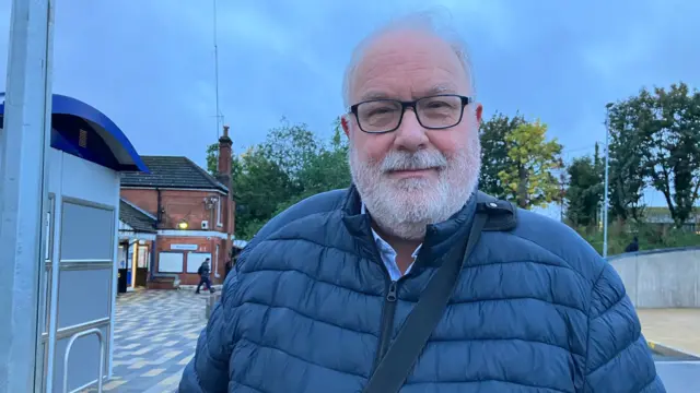 Alan Wiliams, wearing a blue padded jacket and glasses. He has a white beard and is wearing glasses, standing outside Flitwick Railway Station.