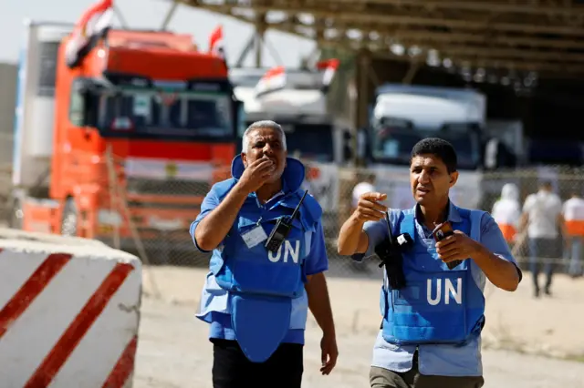 Two men in UN vests stand in front of trucks at the Rafah crossing