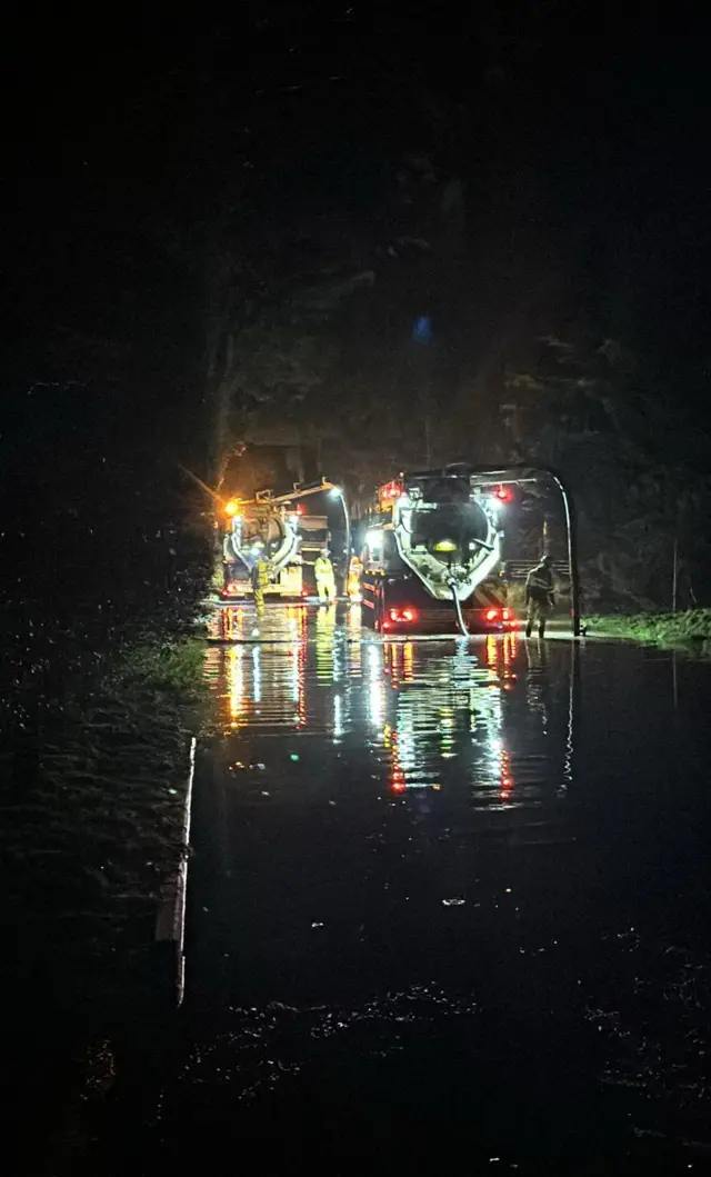 Two tankers in the night can be seen just by their flashing lights pumping water from the road.