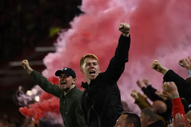 United fans stand and cheer jubilantly as a red flare is let off in the stands