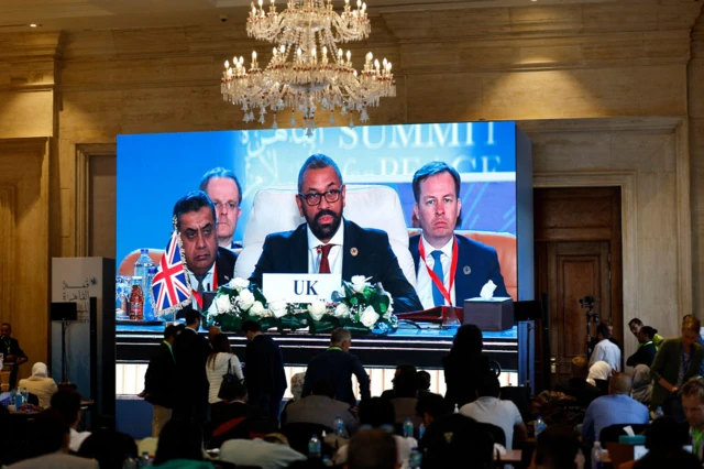 James Cleverly speaks at a summit. His image is televised in front of an audience