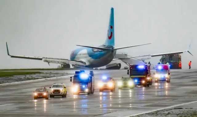 Vehicles on a runway with an aircraft in the background at Leeds Bradford Airport